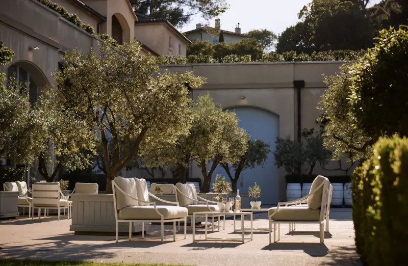 Elegant white lounge chairs and table arranged on sunny Provence château courtyard patio amid olive trees and hedges.