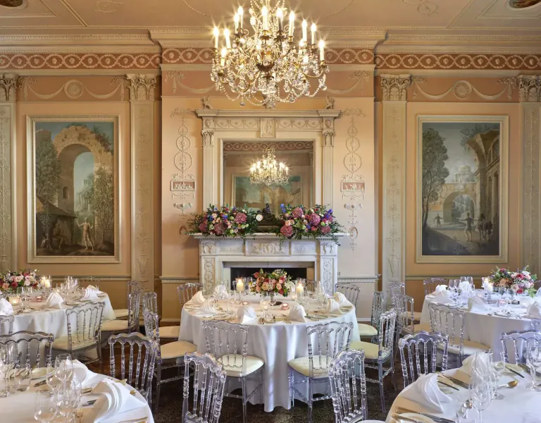 Elegant dining room in Home House club with chandelier, ornate walls, paintings, fireplace, and round tables set with white linens and flowers.