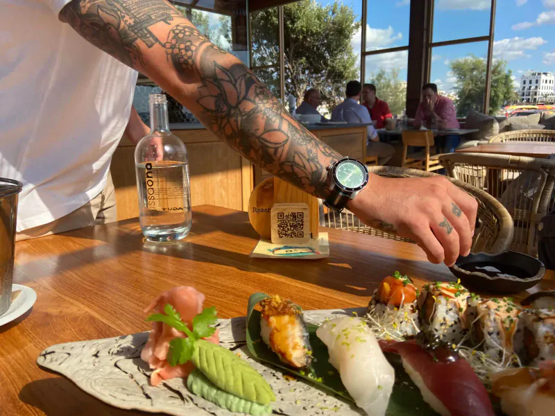 Tattooed hand dipping sushi into soy sauce at Assaona Gastrobeach Palma Club, with water bottle and QR code stand.