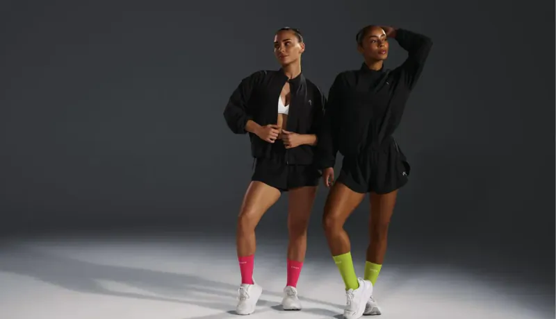 Two women in black Oner Active hybrid outfits with pink and green socks pose confidently on dark studio background.