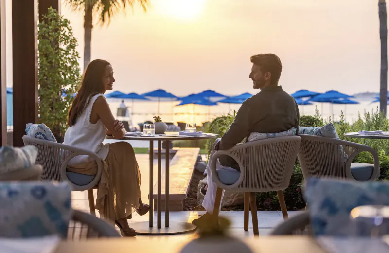Couple seated at outdoor table conversing at sunset, Four Seasons Resort Dubai Sea Fu, blue umbrellas and sea view.