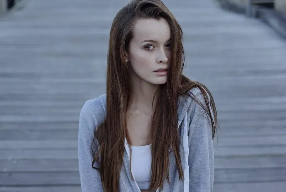 Young woman with long straight brown hair wearing gray hoodie and white tank top on wooden pier