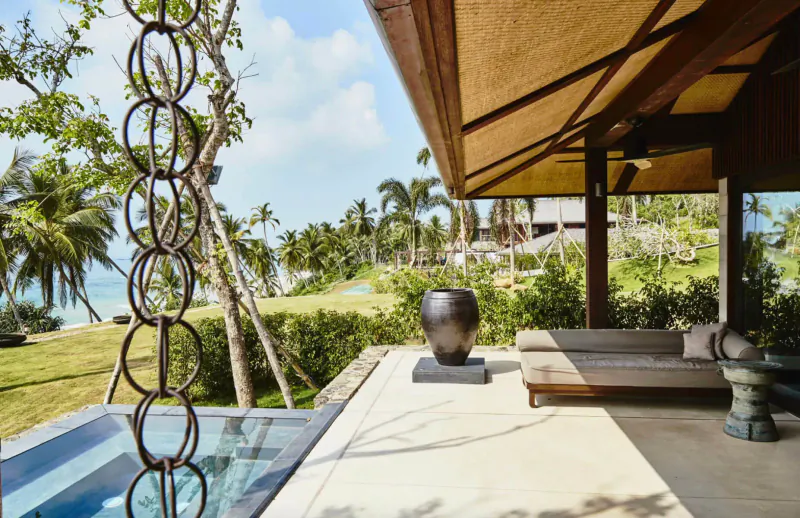 Rustic wooden private resort deck with chain fountain, large vase, daybed, overlooking tropical beach and palms