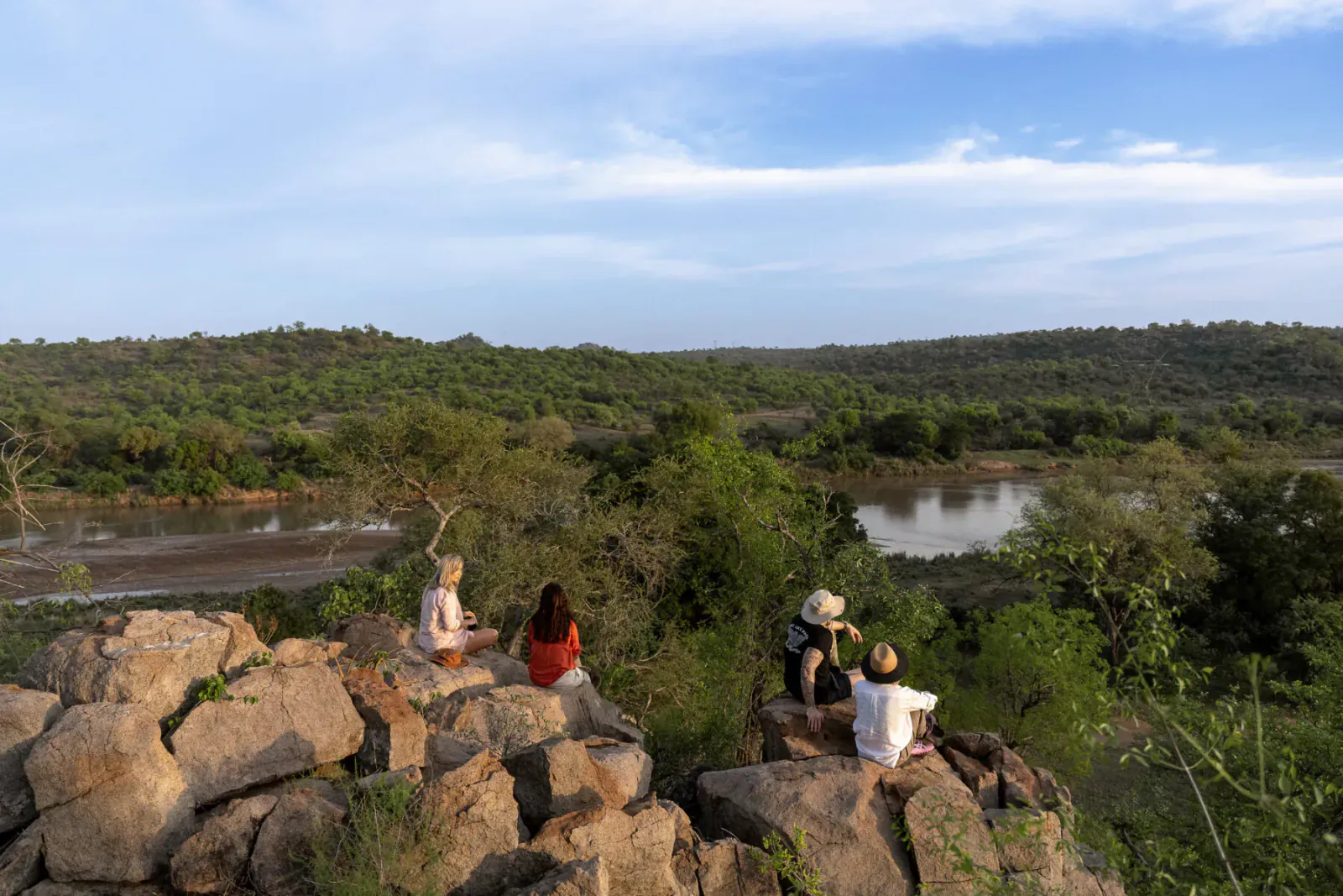 Group of people in hats and casual clothes sitting on rocks overlooking lush green hills, river, and blue sky at Sashwa River of Stars.