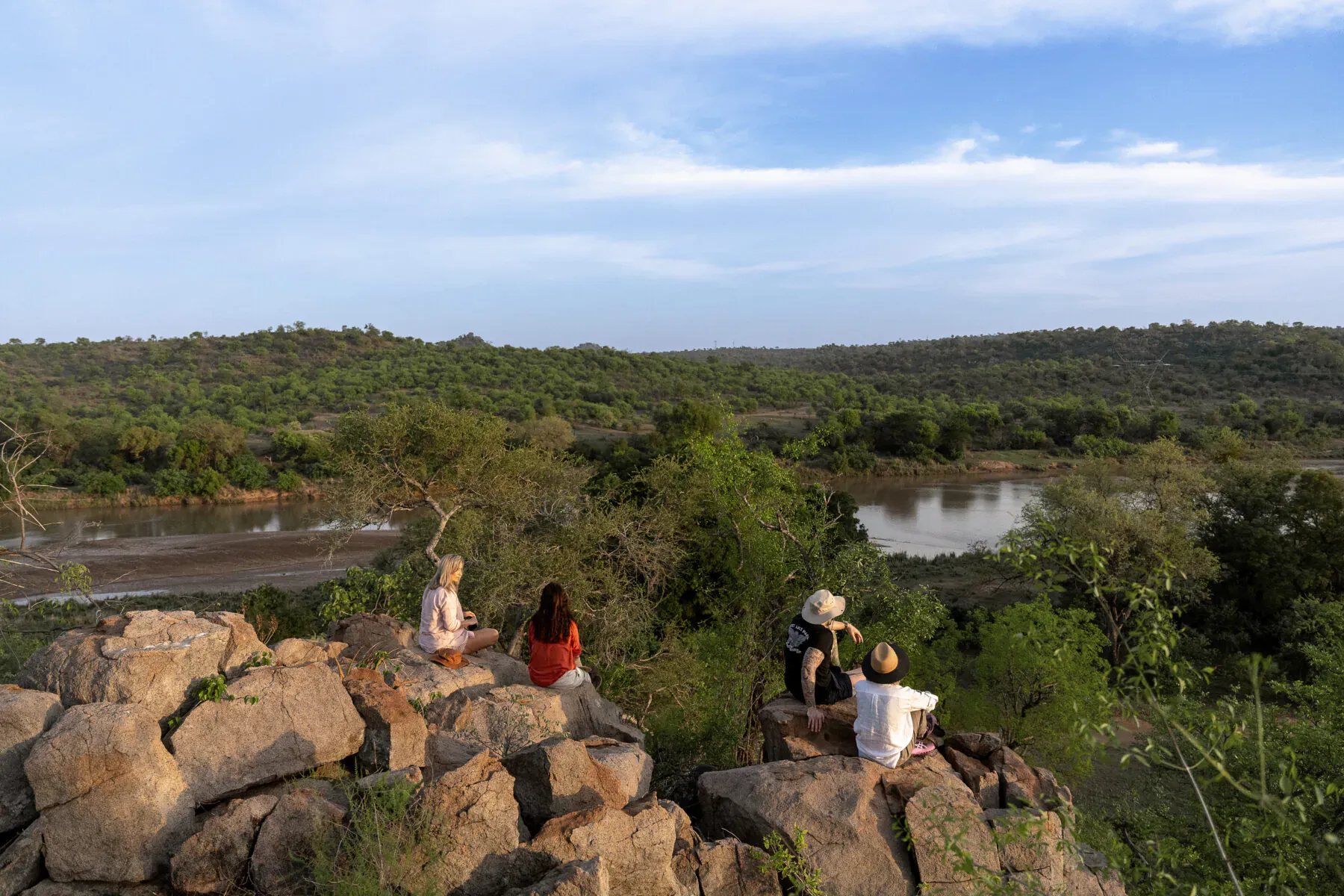 Group of people in hats and casual clothes sitting on rocks overlooking lush green hills, river, and blue sky at Sashwa River of Stars.