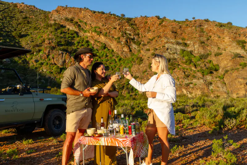 Three people toasting with wine glasses beside a table of drinks and snacks next to a safari vehicle at Mont Eco Game Reserve, rocky hills backdrop.