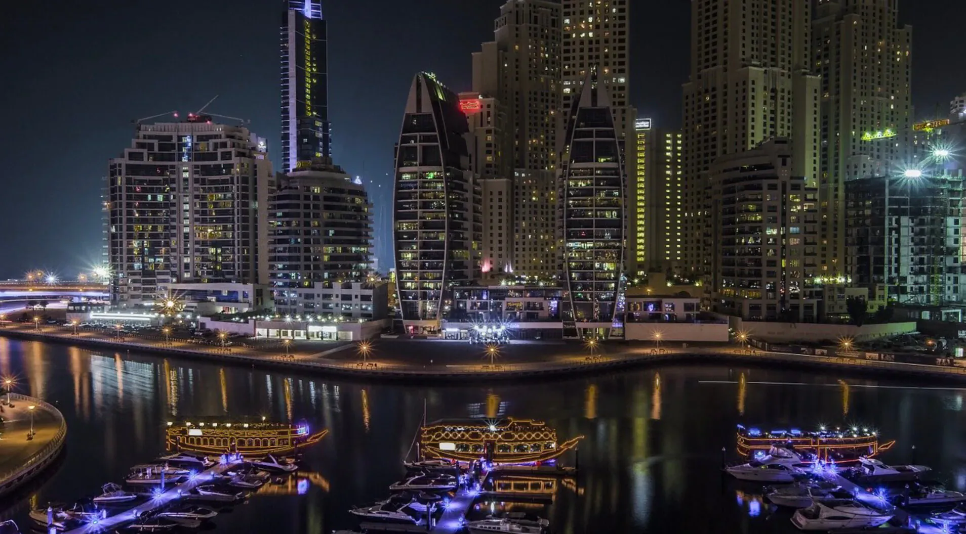 Nighttime Dubai skyline with glittering skyscrapers and lit luxury yachts on the waterfront.