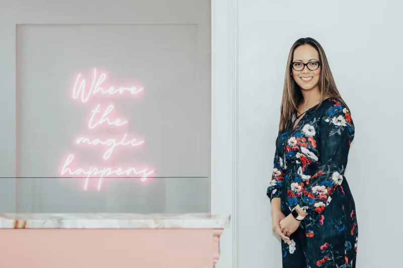 Woman in floral navy dress stands by 'Where the magic happens' neon sign at stylish clinic reception.