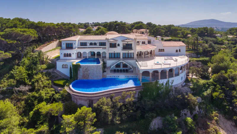Aerial view of luxurious white villa with infinity pool on a lush green hillside overlooking mountains.