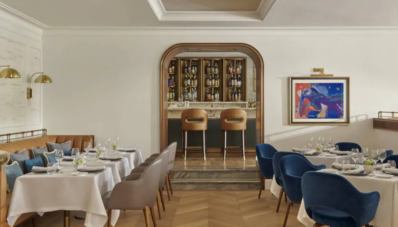 Elegant hotel dining area with white walls, parquet floor, blue and gray upholstered chairs at set tables, wooden bar stools, and abstract art.