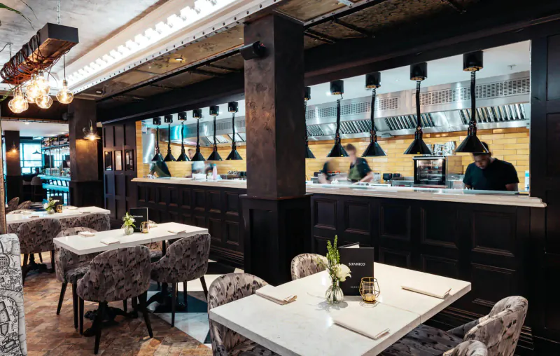 Elegant Six by Nico restaurant interior in Canary Wharf with Ancient Rome theme, dark wood bar, chefs at open kitchen, set tables with flowers.