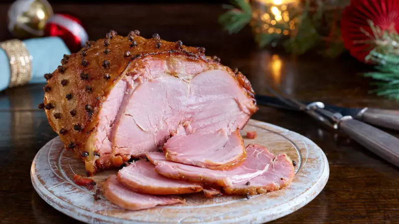 Sliced glazed ham with cloves on a plate, garnished with Christmas ornaments, pine, and candles.