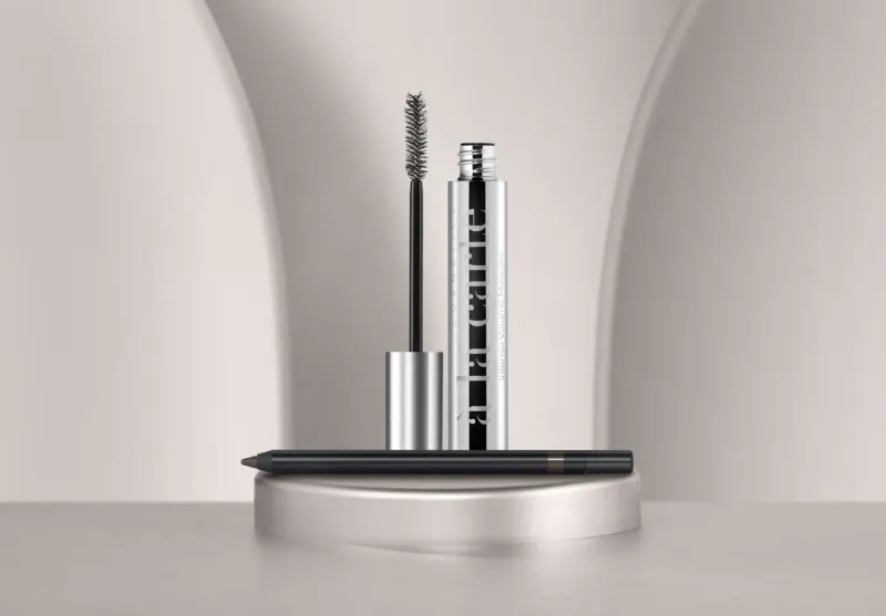 Silver Diptyque mascara tube with brush and black eyeliner pencil on round stand against curved backdrop