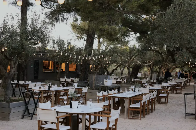 Outdoor dining area at Château Léoube Provence with wooden tables, white chairs, string lights, olive and pine trees on sandy ground.