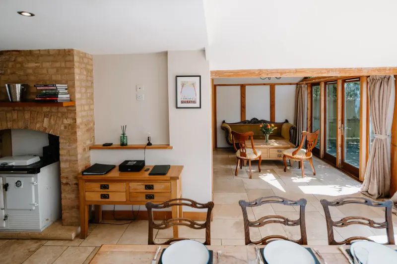 Elegant Cotswolds country house kitchen and dining area with brick fireplace, wooden beams, and sunlit windows.