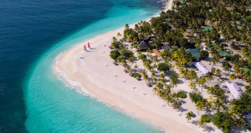 Aerial view of Palm Island Resort & Spa: white sand beach curving into turquoise waters, red sails, palm trees, and bungalows.