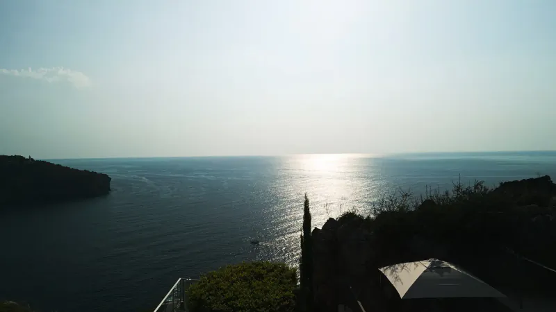 Scenic sunset over the sea from a cliffside terrace with greenery and shaded canopy at Es Fanals restaurant