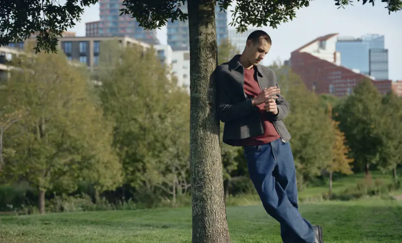 Man in gray jacket and blue jeans leans against tree, looking at phone, with Canary Wharf skyscrapers and green park behind.