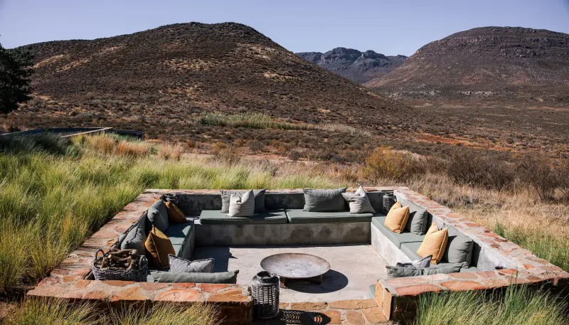 Outdoor stone-walled lounge with green and yellow cushions around a fire pit, set in grassy fynbos with rugged mountains beyond, Simbavati Cederberg Ridge.
