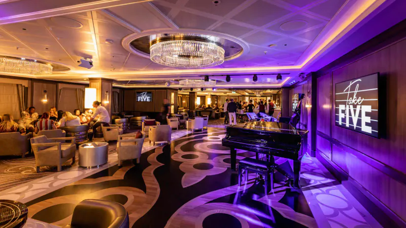 Elegant Princess Cruises 'FIVE' lounge with grand chandelier, grand piano, plush seating, and patrons under purple lighting.