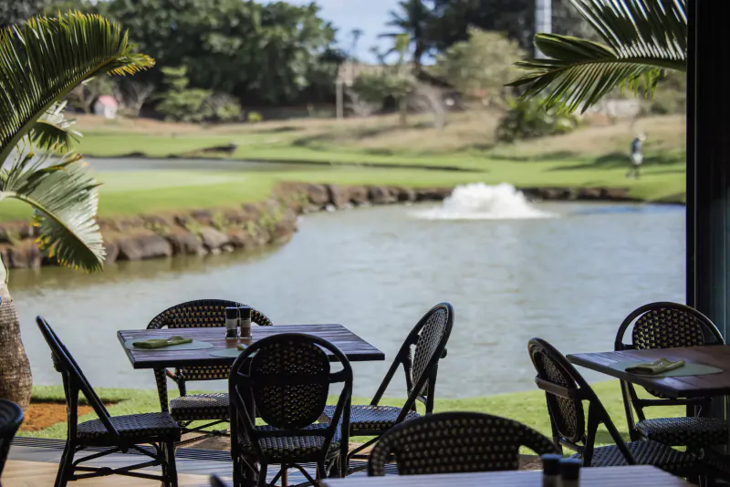 Outdoor terrace at Mauritius golf course with wicker tables set for dining, overlooking lush green fairway, water hazard with fountain, and palm trees.