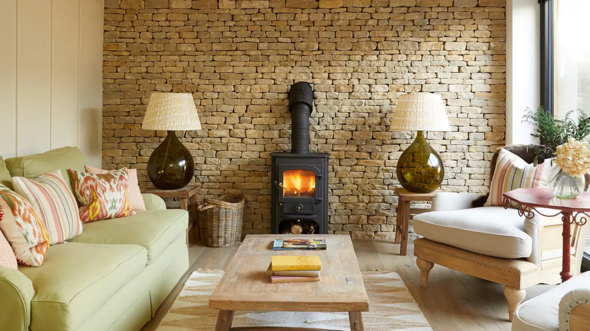 Cozy living room in Kingham Plough Little Barn with green sofas, lit wood-burning stove, green glass lamps, and flowers on stone wall.