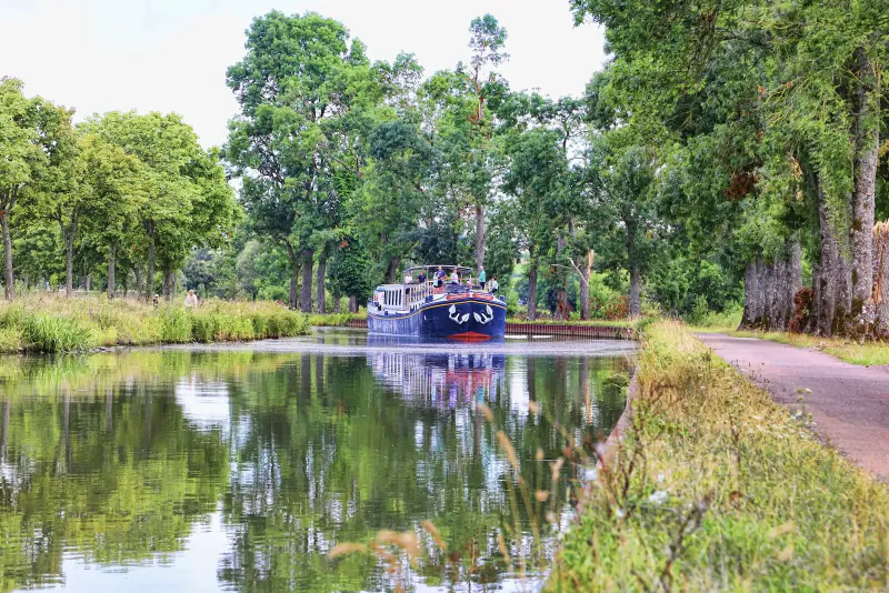 Dark blue barge cruising through tree-lined canal in Burgundy wine country, with clear reflections in calm water.