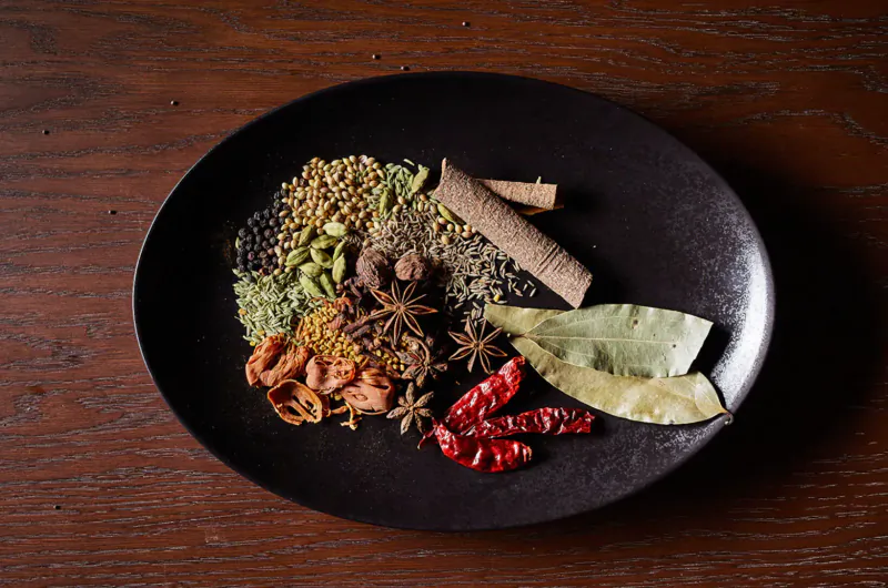 Assortment of Indian spices on black plate: black pepper, cumin, cardamom, star anise, cinnamon, bay leaves, dried chilies.