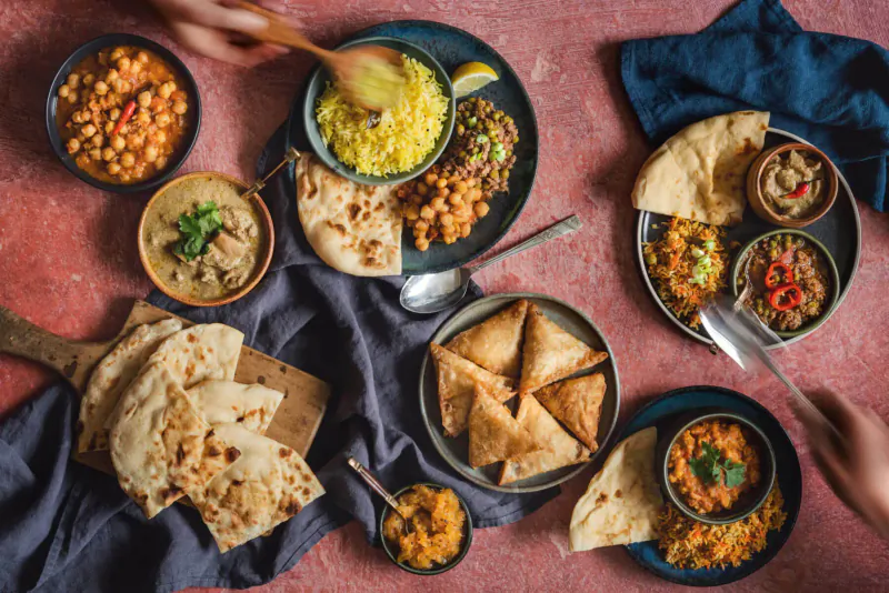 Assortment of Indian dishes on pink table: chickpeas, yellow rice, naan, samosas, curries, hands serving with spoons.