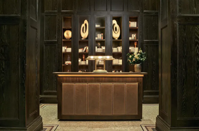 Elegant wooden reception desk in grand dark-paneled library lounge at Kimpton Fitzroy London, with bookshelves, lamp, and flowers.