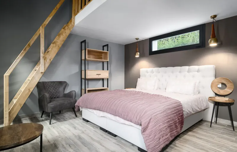 Cozy loft bedroom with white tufted bed, pink bedding, wooden staircase, bookshelves, armchair, and window overlooking greenery.