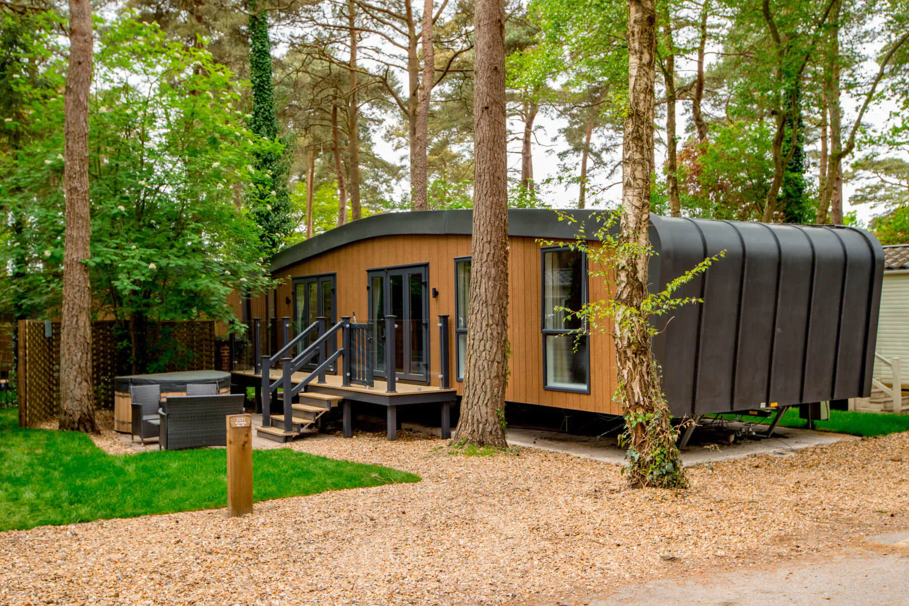 Modern wooden cabin with curved black roof at Sandy Balls Holiday Village in New Forest, surrounded by tall trees, deck, and lawn.