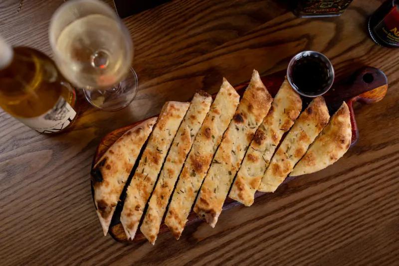 Sliced garlic flatbread on wooden board with balsamic dip, white wine glass, and beer bottle on table