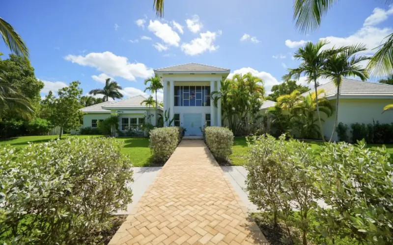 Ciao Bella luxury estate in Lyford Cay, white mansion with blue windows, palm trees, brick pathway, lush gardens under blue sky