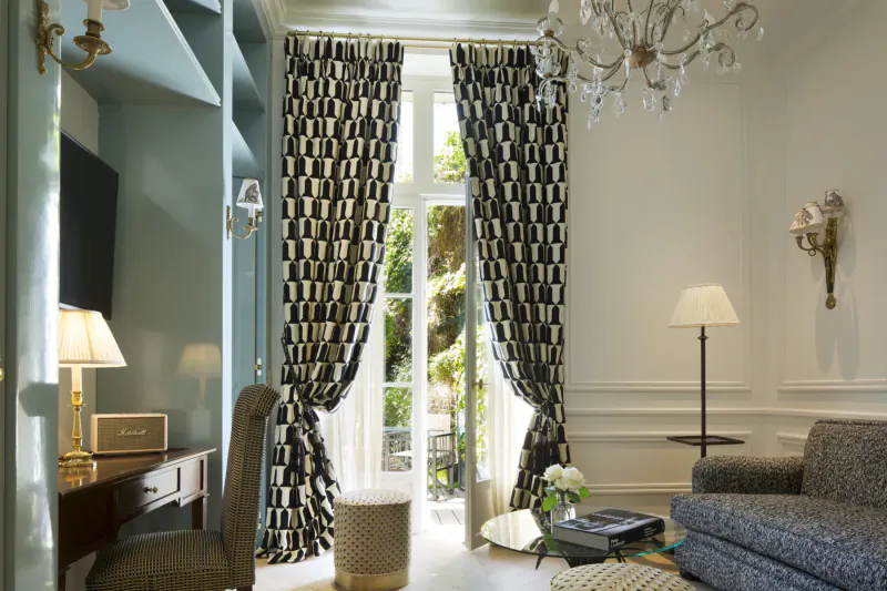 Elegant hotel room at Relais Christine Paris with geometric black-and-white curtains framing French doors to garden, chandelier, sofa, desk, and lamps.
