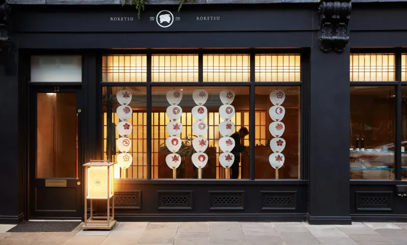 Roketsu restaurant facade at night with warm-lit windows displaying white and red circular art, lantern outside, 'Roketsu' sign.