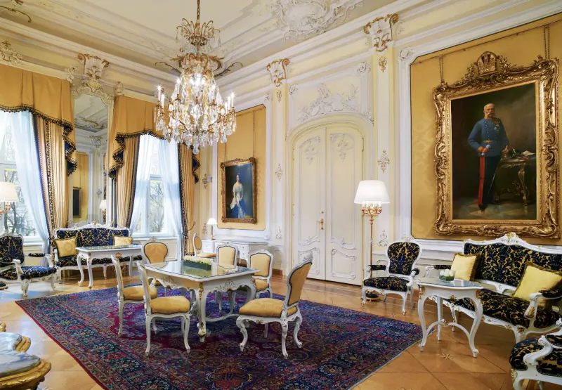 Opulent rococo room in Hotel Imperial Vienna with chandelier, gilded portraits of Emperor Franz Joseph, ornate furniture, and velvet drapes.