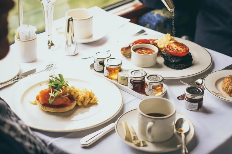 Elegant English breakfast on train dining table: smoked salmon muffin, eggs Benedict, tea, jams, with green view outside.
