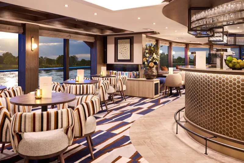 Elegant river cruise dining lounge with curved bar, striped chairs, sunset river view, and potted plants.