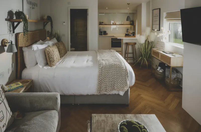 Cozy studio retreat with white bed, plush pillows, plants, kitchenette, TV, and wooden floors at Everything Retreat.