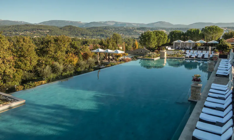 Aerial view of infinity pool with loungers at Terre Blanche Hotel Spa Golf Resort in Provence, surrounded by hills and trees.