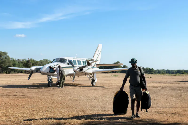 Man in green hat carrying black bags walks toward white twin-engine plane on Zambian bush airstrip with crew nearby.
