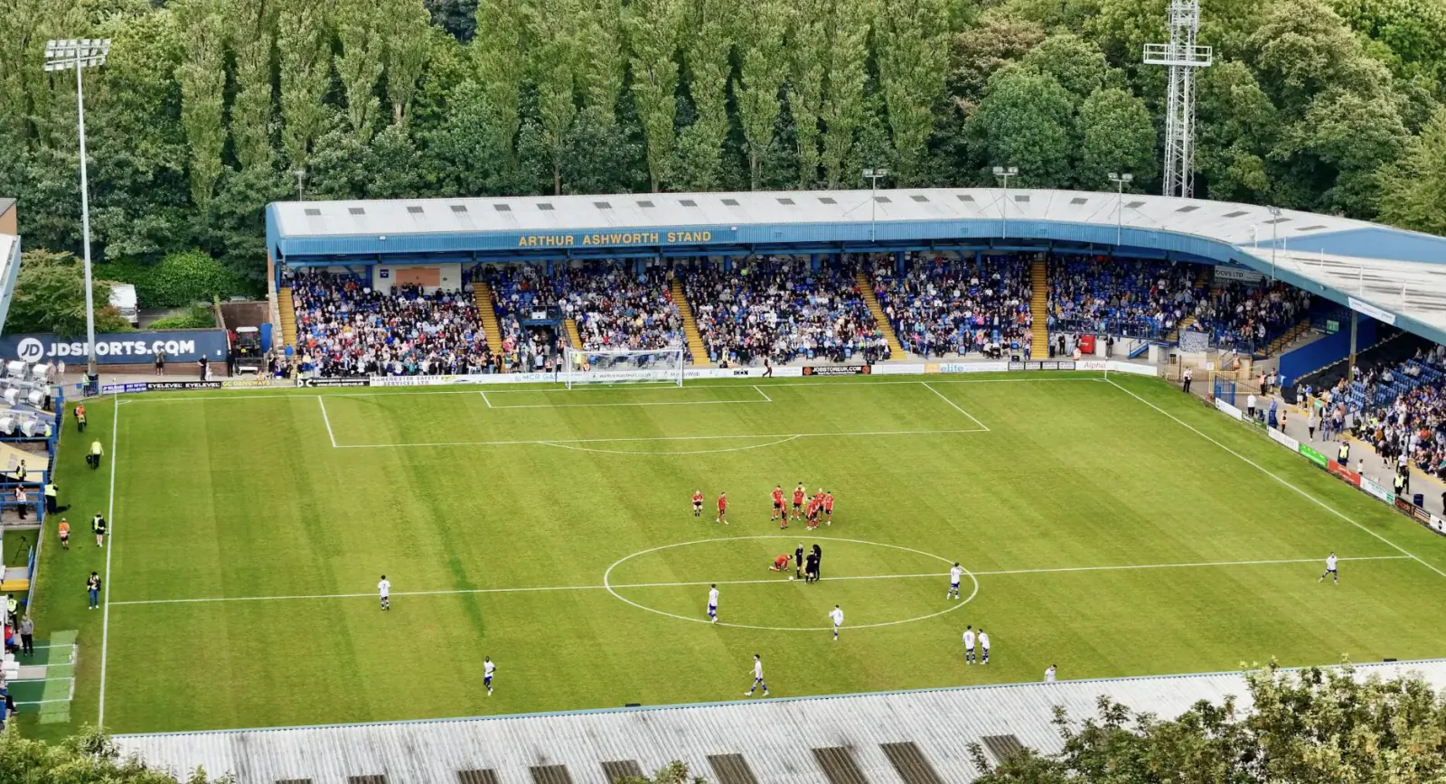 Aerial view of a soccer stadium with blue stands full of fans, green pitch, and players in red and white during a match, surrounded by trees.