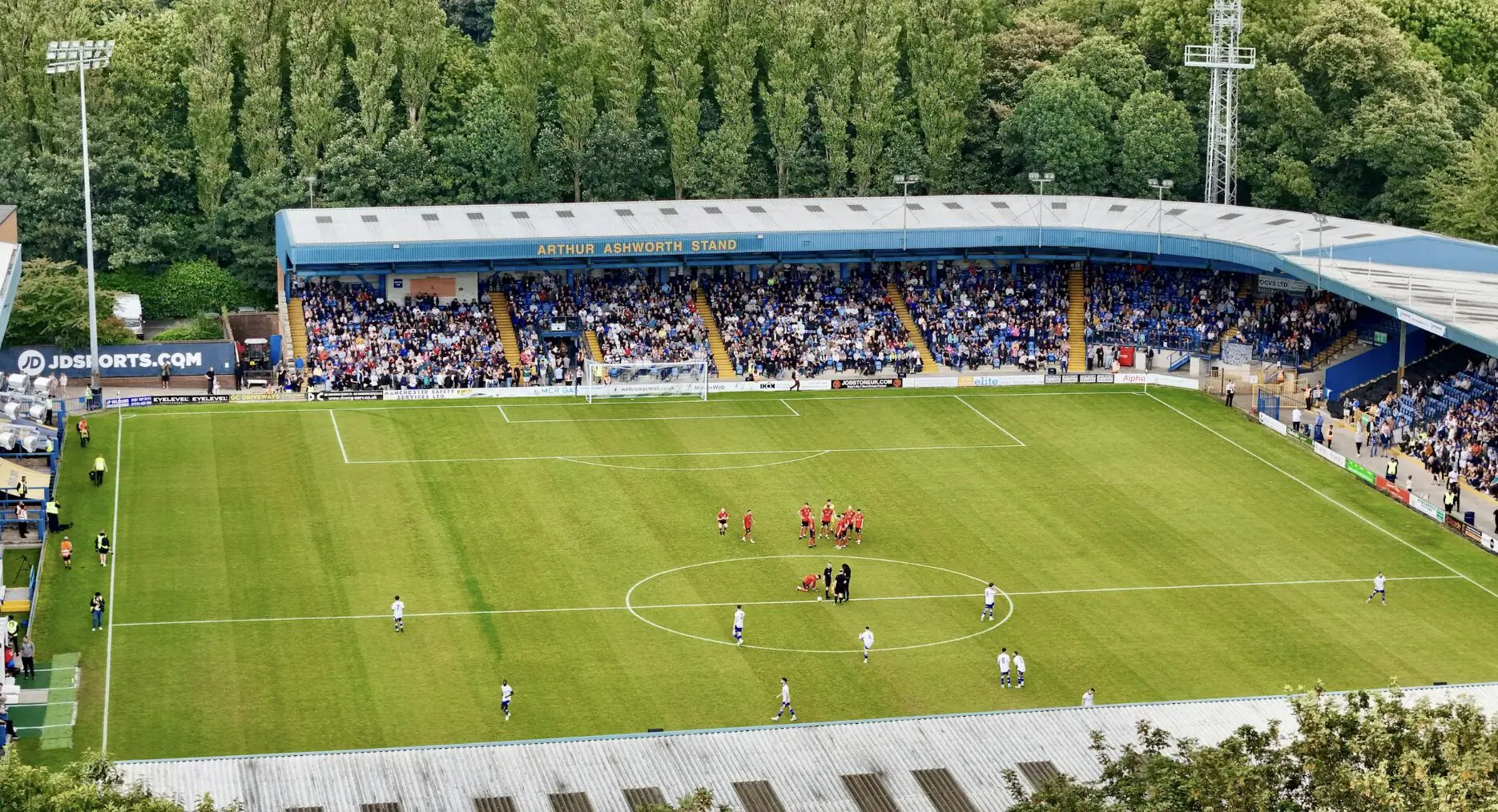 Aerial view of a soccer stadium with blue stands full of fans, green pitch, and players in red and white during a match, surrounded by trees.