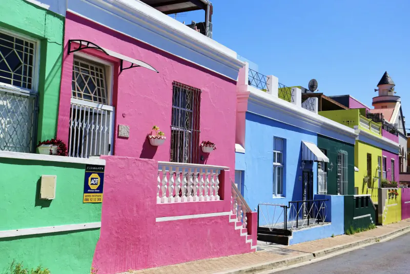Colorful Cape Malay houses in vibrant pinks, greens, blues along sunny street with church steeple.