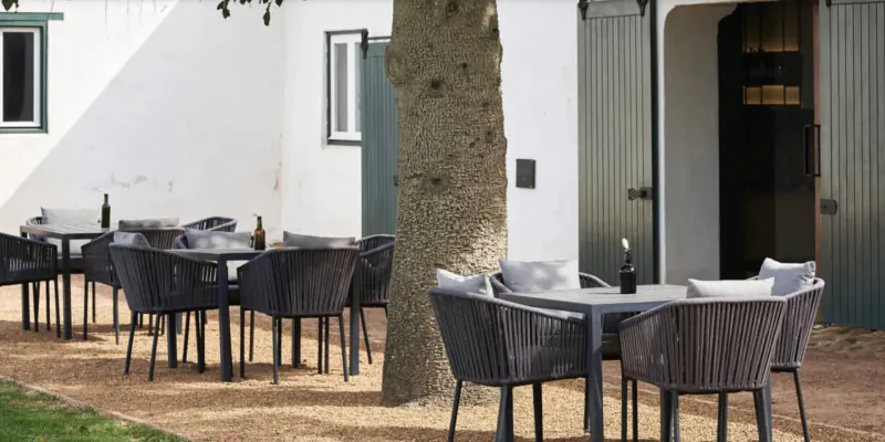 Outdoor seating area at Clara’s Barn, Vergenoegd Löw wine estate, with wicker tables, chairs, bottles under a tree by white building