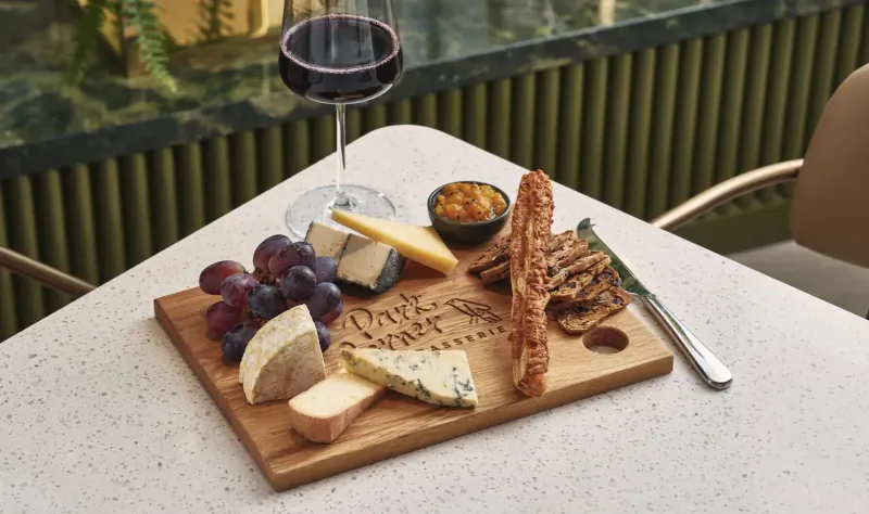 Park Corner Brasserie charcuterie board with cheeses, grapes, chutney, crackers on wooden board, red wine glass beside, green marble backdrop.