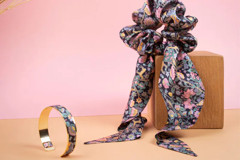 Large navy floral scrunchie draped over wooden block with matching gold cuff bracelet on pinkish surface
