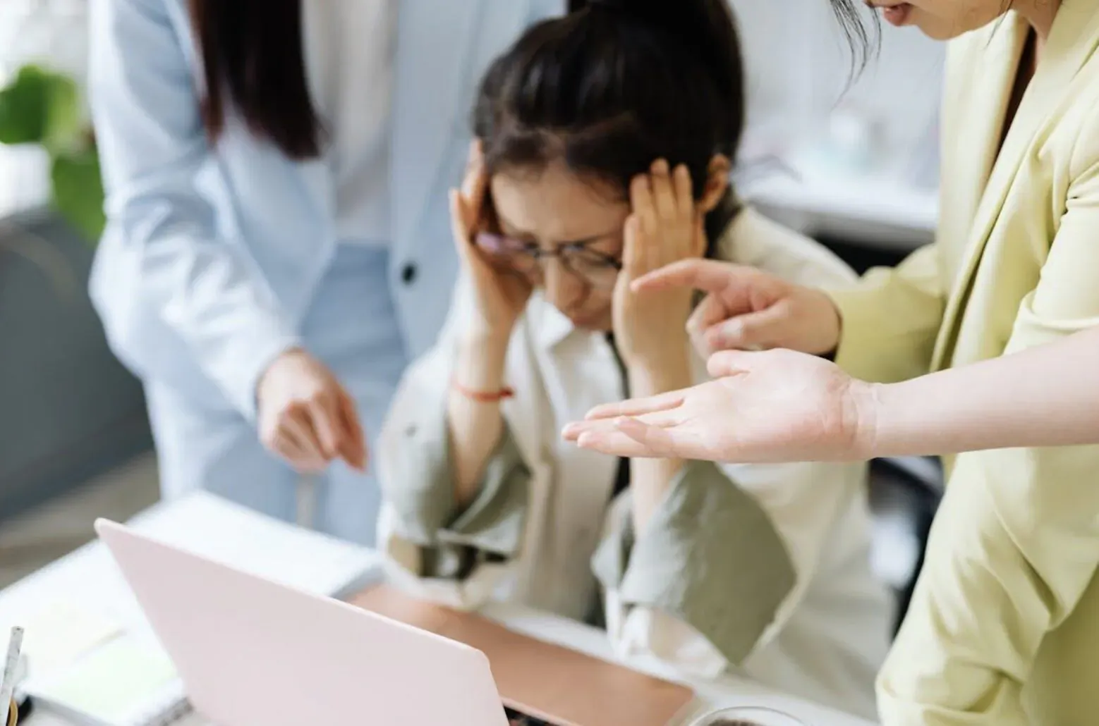 Stressed Asian woman in glasses holding head at laptop, colleagues in light suits pointing and gesturing around desk.