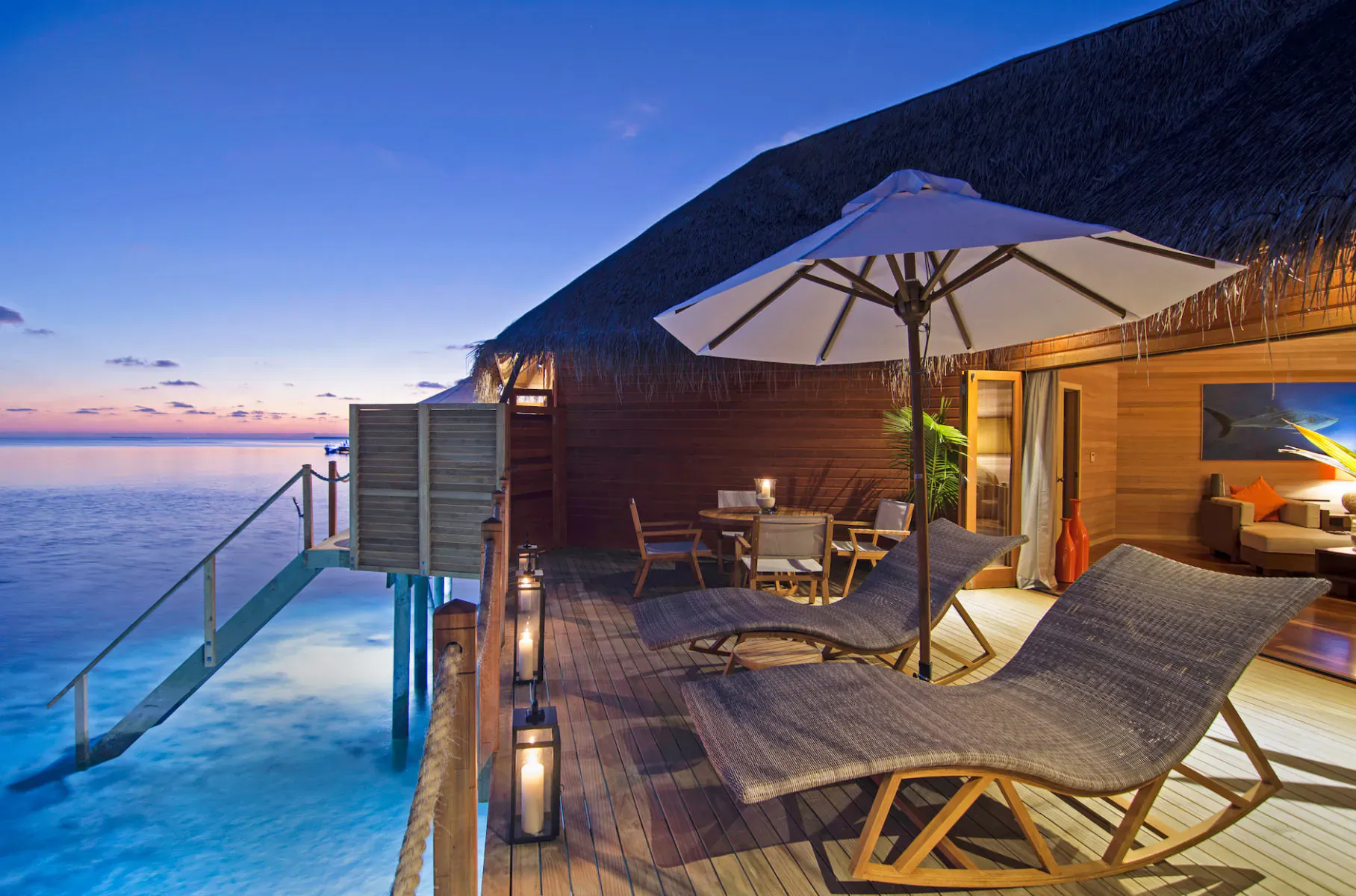 Twilight view of Mirihi Island Resort overwater villa deck with loungers, table, lanterns, and turquoise lagoon.